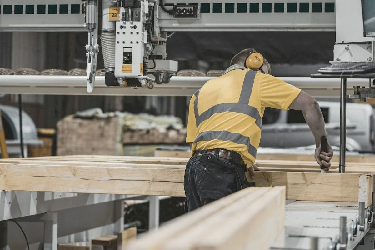 Een medewerker in werkkleding met gehoorbescherming verwerkt houten balken in een productiehal