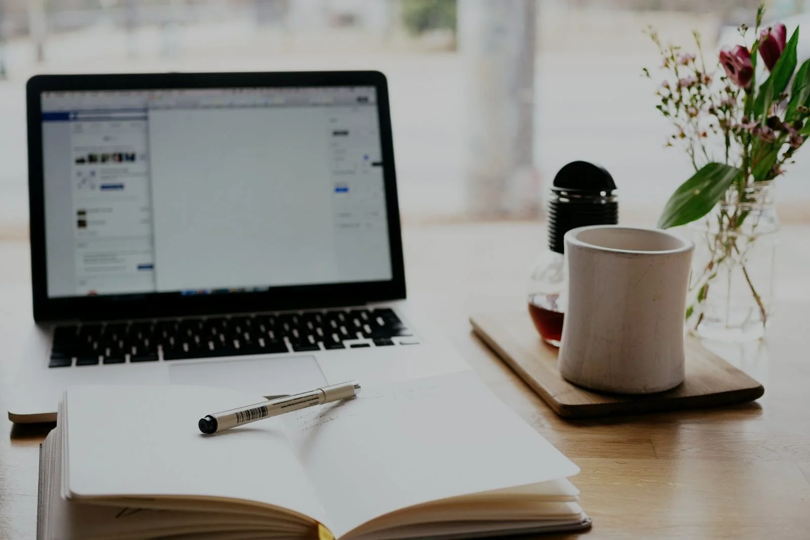 Een laptop, een opengeslagen notitieboek met pen en een mok op een houten tafel bij het raam