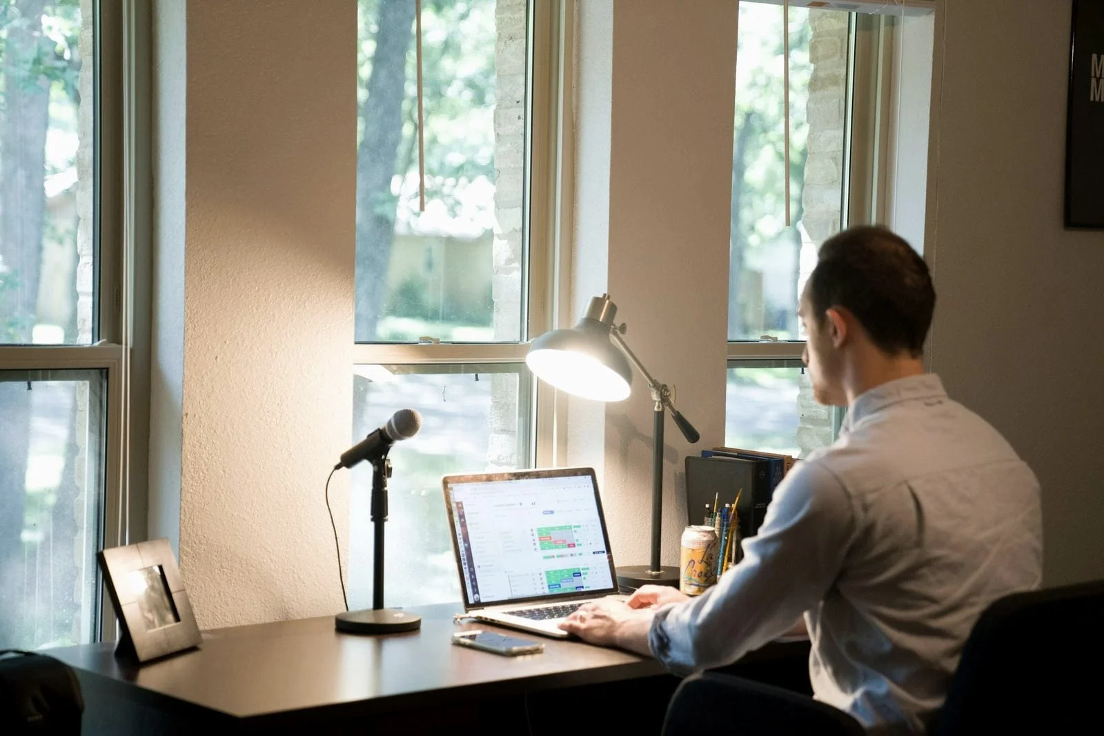 Een man werkt thuis op zijn laptop aan een bureau bij het raam, met een bureaulamp en microfoon