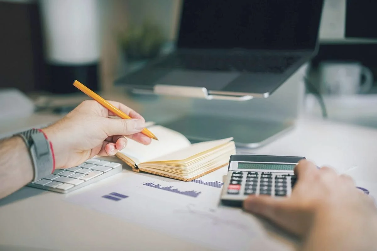 Iemand werkt de administratie bij met rekenmachine, notitieboek en overzichten op het bureau