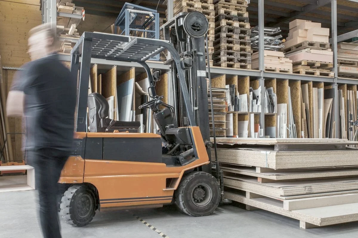 Een medewerker loopt langs een heftruck en stellingen met pallets in een magazijn