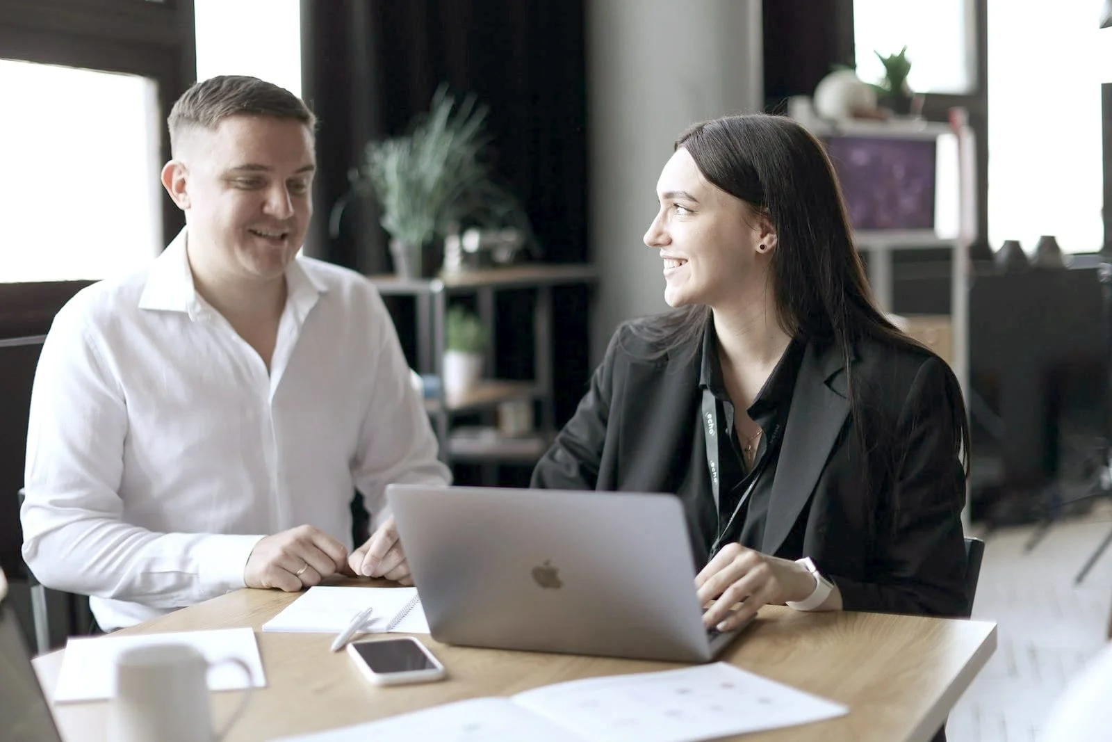 Twee mensen voeren een ontspannen kennismakingsgesprek aan tafel, met een laptop en notities tussen hen in