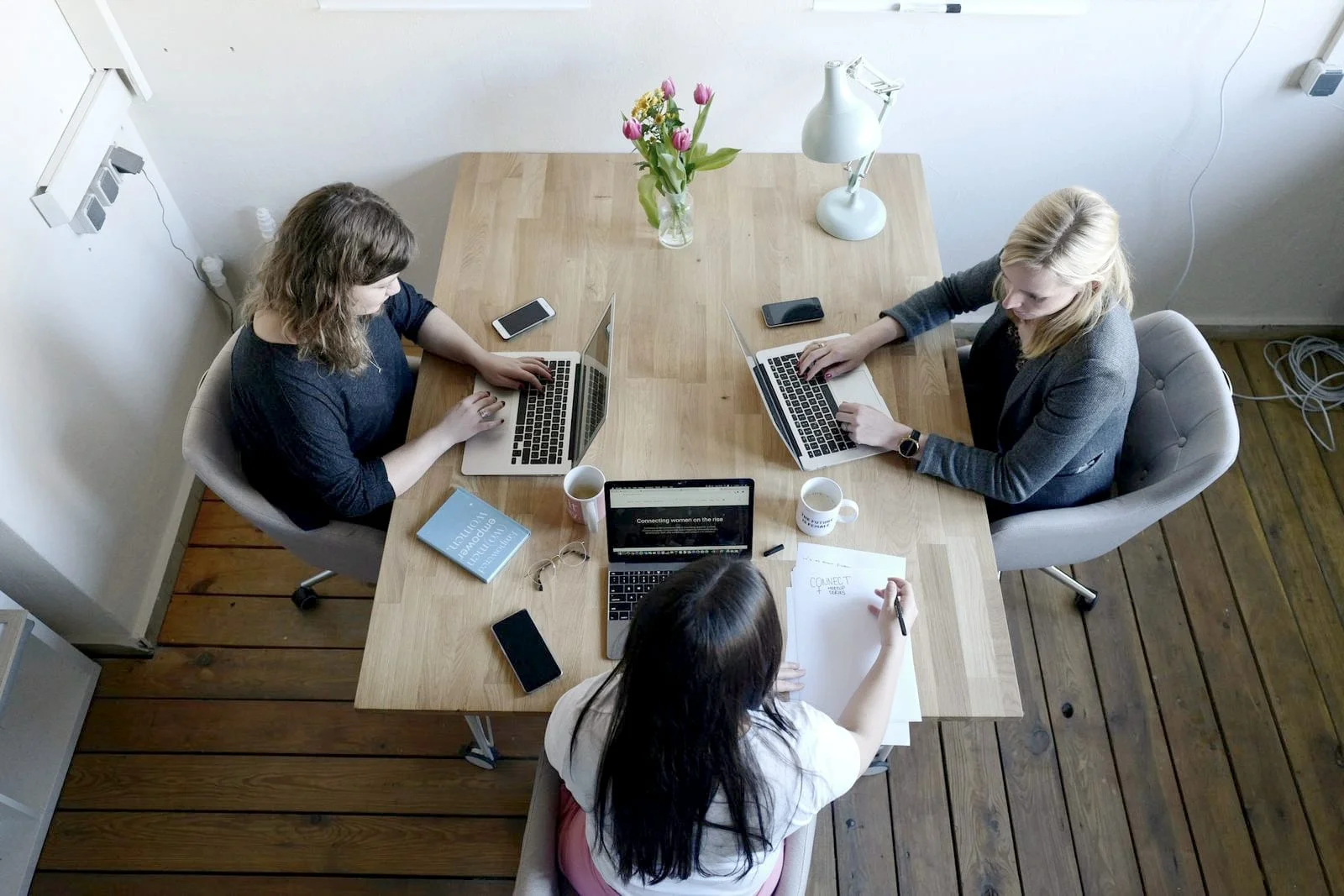 Drie vrouwen werken samen aan een houten tafel met laptops en notities, van bovenaf gezien